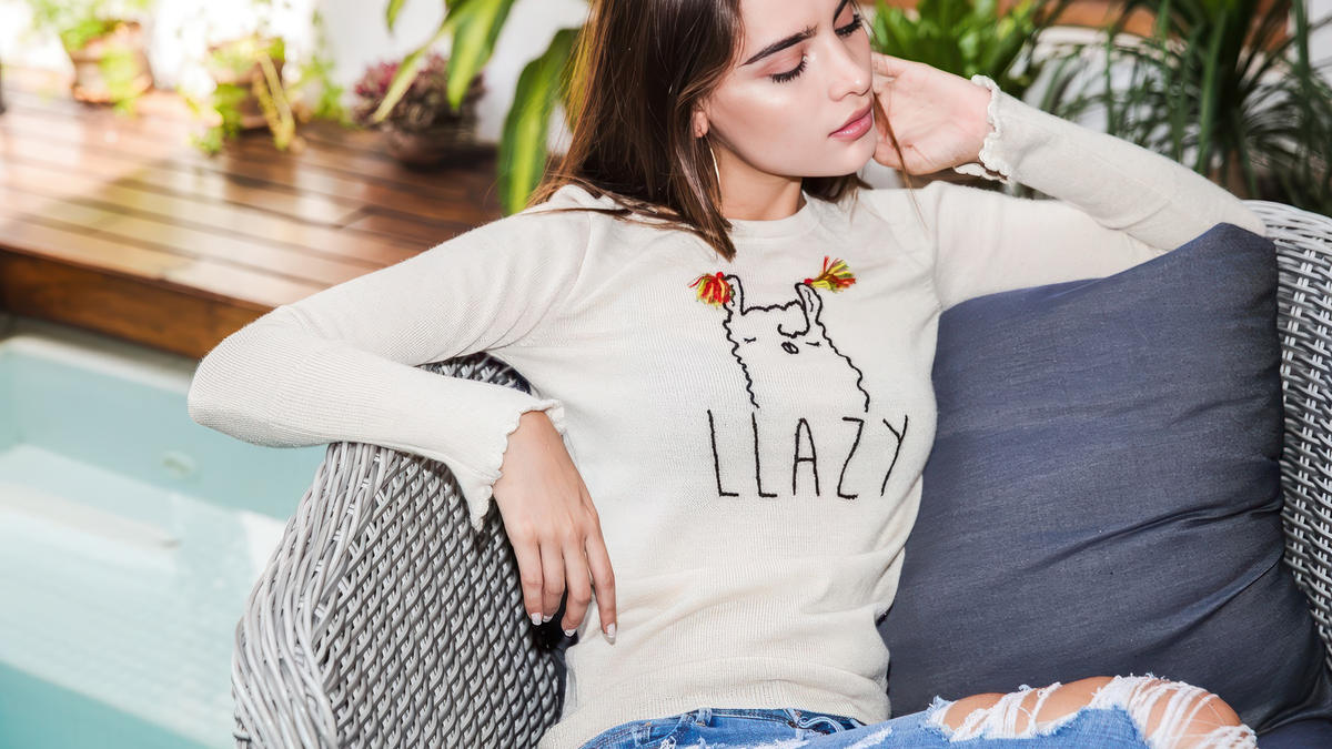 Woman relaxing in a chair wearing a cream alpaca sweater with embroidered llama and text LLAZY, part of Yapa Alpaca Store collection.